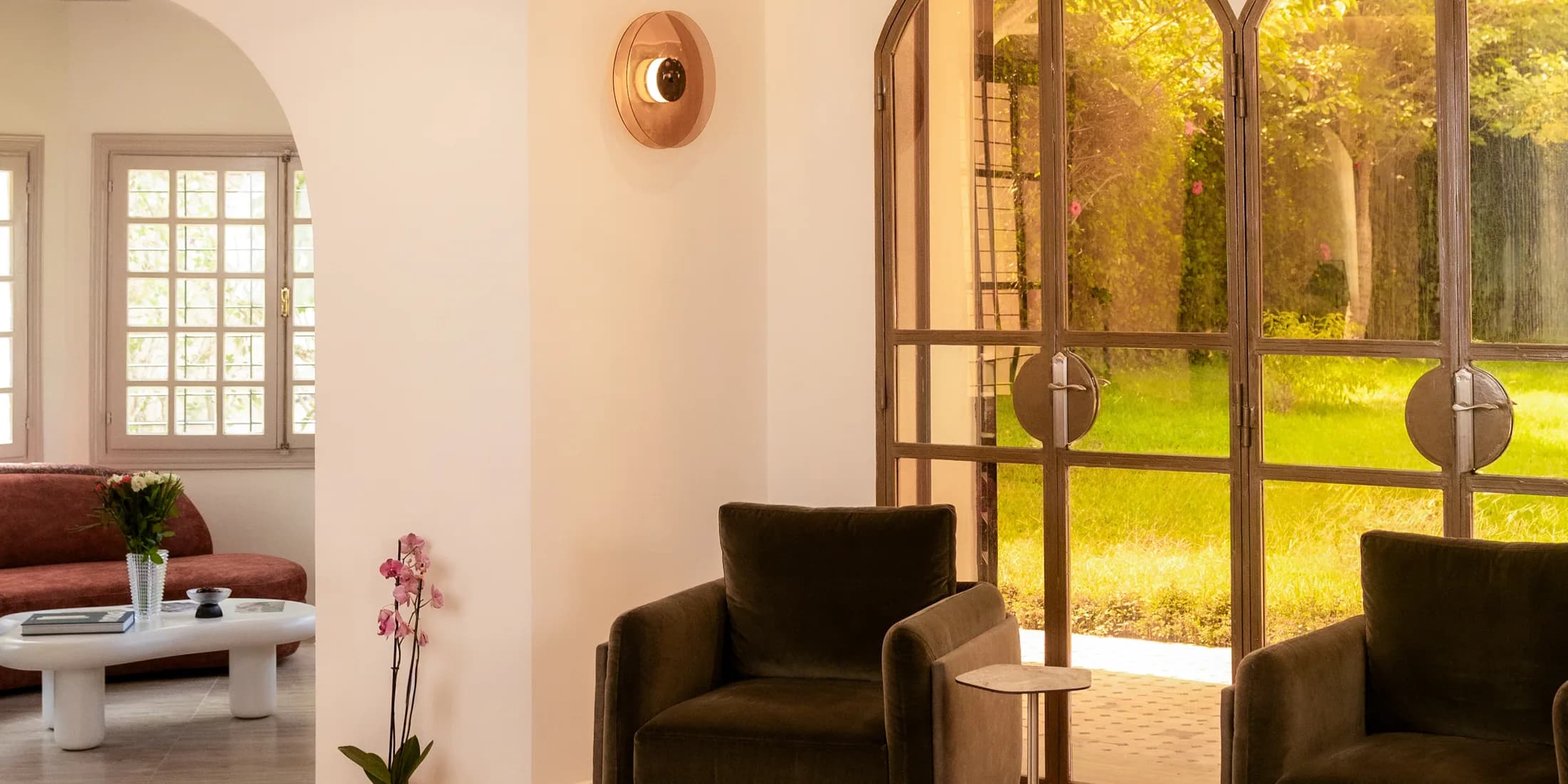 A clinic waiting lounge with velvet armchairs, an orchid and a low table, beside tall arched doors standing open onto a sunlit garden.